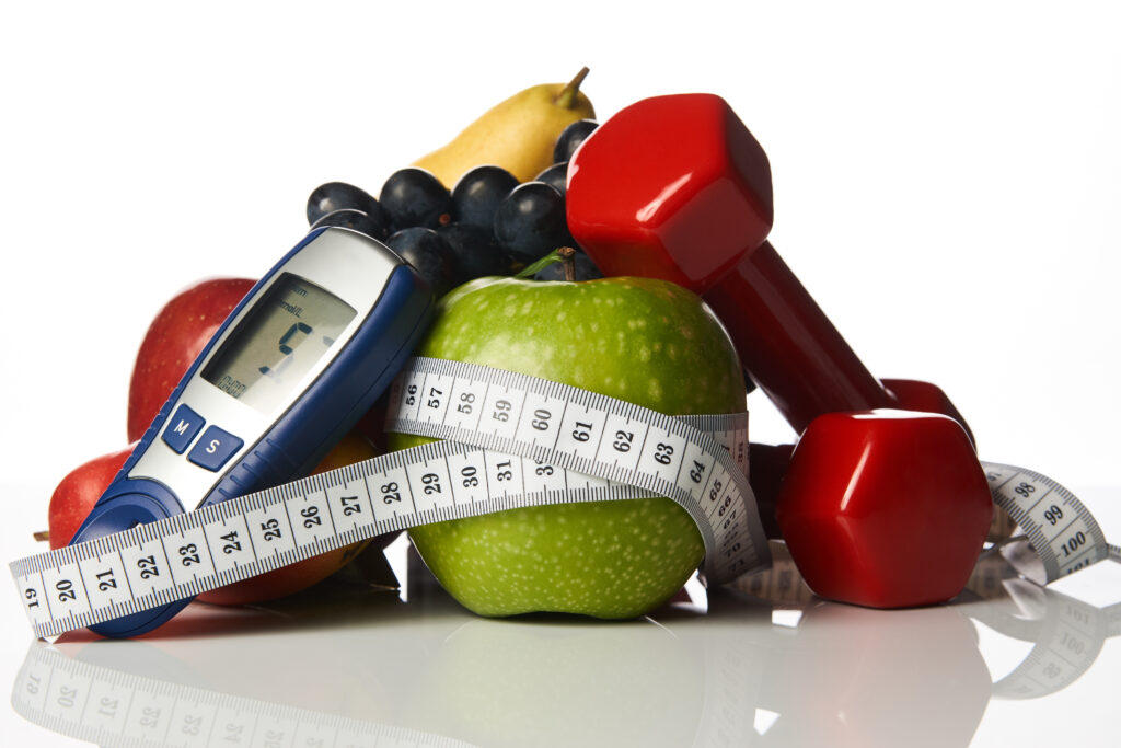 Blood sugar control glucometer for glucose level with healthy organic food and dumbbells with measuring tape on a white background.