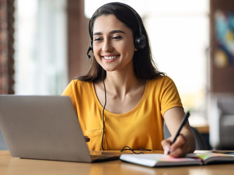Happy young arab woman having educational course online, using laptop and headset, cafe interior, free space. Smiling female student studying online, sitting in front of laptop at cafeteria