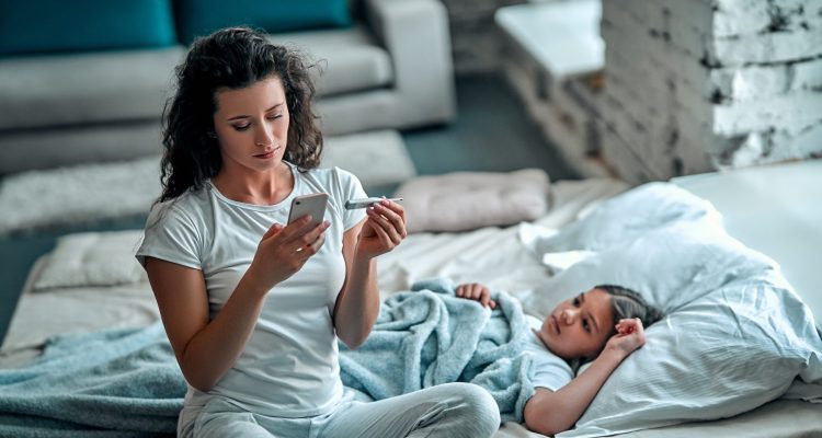 Mother measuring temperature of her ill kid. Sick child with high fever laying in bed and mother holding thermometer. Mother with cell phone calling to doctor