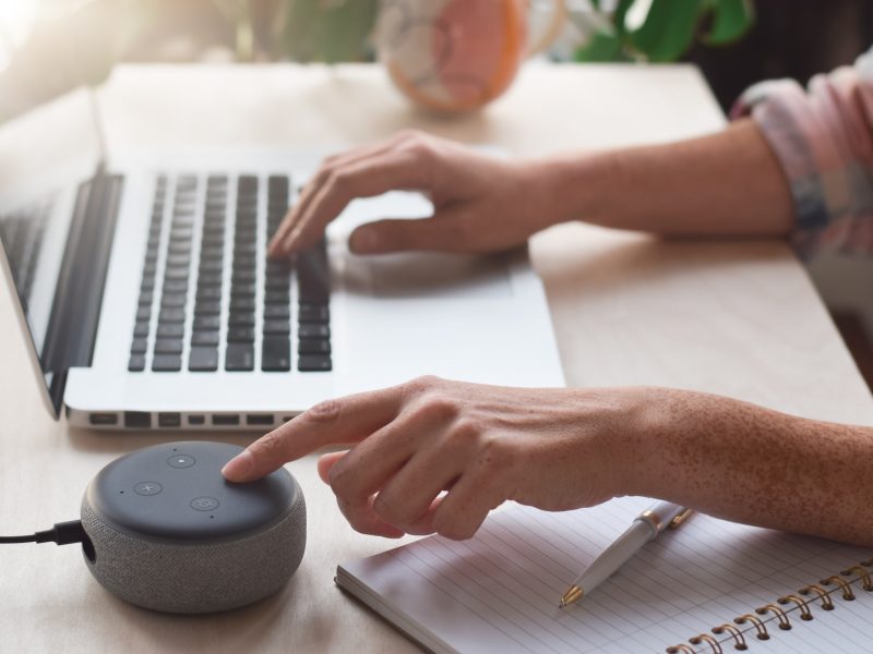 woman-sitting-at-desk-using-laptop-computer-and-pressing-button-on-virtual-assistant-smart-speaker.jpg